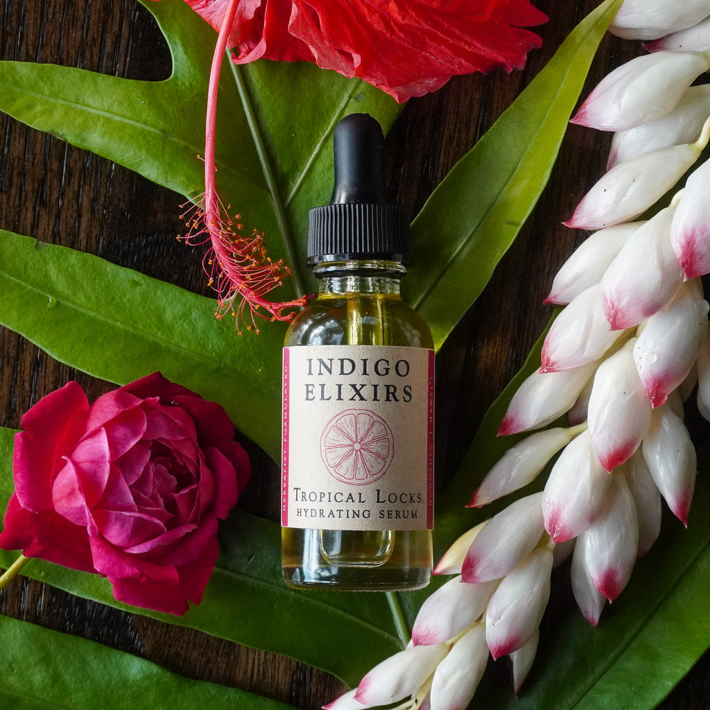 Indigo Elixirs Tropical Locks Hydrating Serum bottle surrounded by flowers on a wooden surface