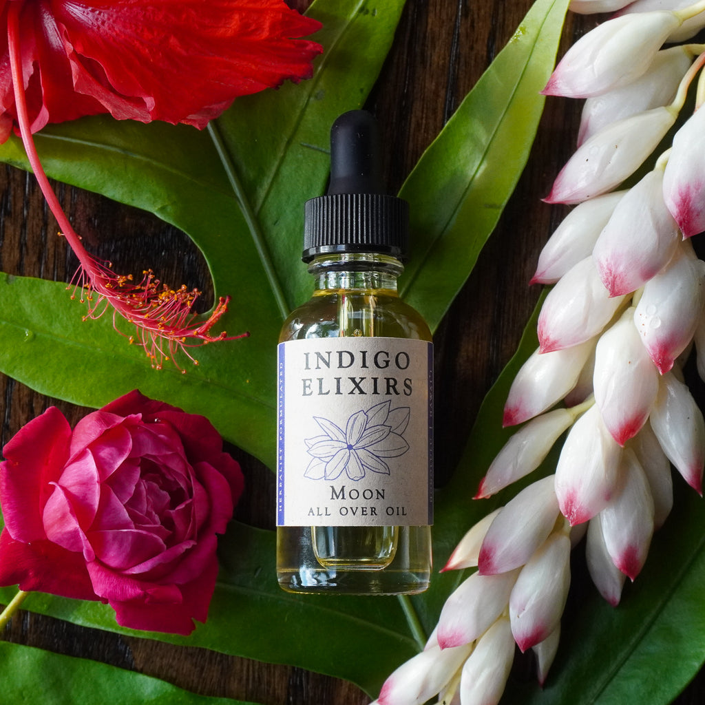 Bottle of Indigo Elixirs Moon All Over Oil surrounded by tropical flowers on a wooden surface