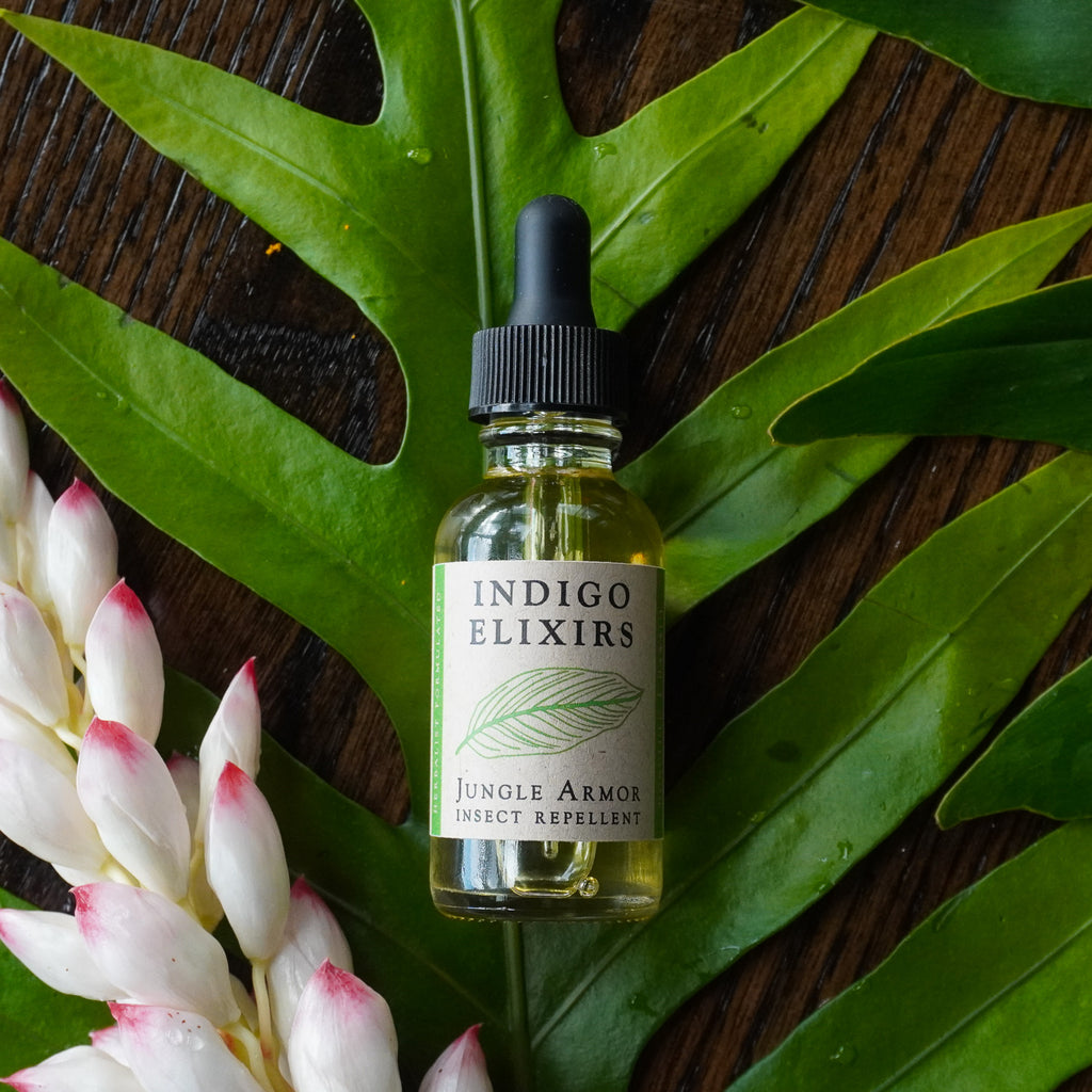 Bottle of Indigo Elixirs Jungle Armor insect repellent on green leaves with pink flowers.